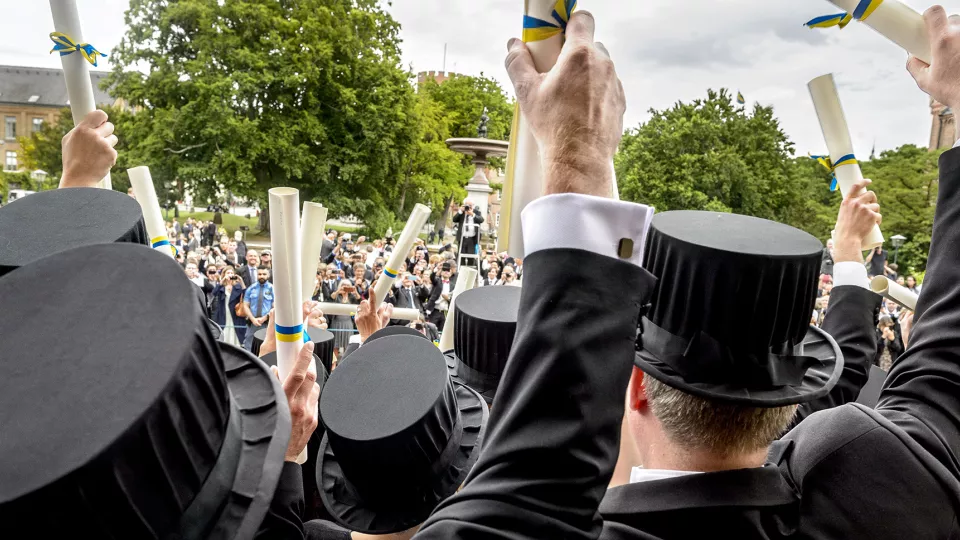 Personer i höga svarta hattar vinkar med ihoprullade diplom. Foto.