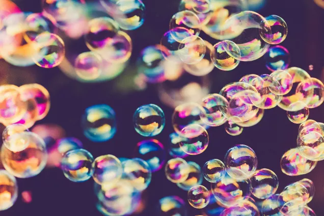 Bubblor i regnbågsfärger mot mörk bagrund. Foto från Unsplash.