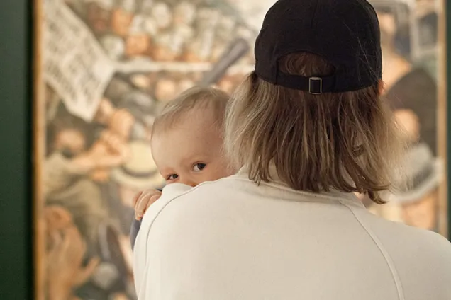 10 Visning 250522 Foto Emma Krantz.jpg Ett litet barn kikar fram bakom en förälders axel och tittar in i kameran. Föräldern har ryggen mot kameran och tittar på en målning.