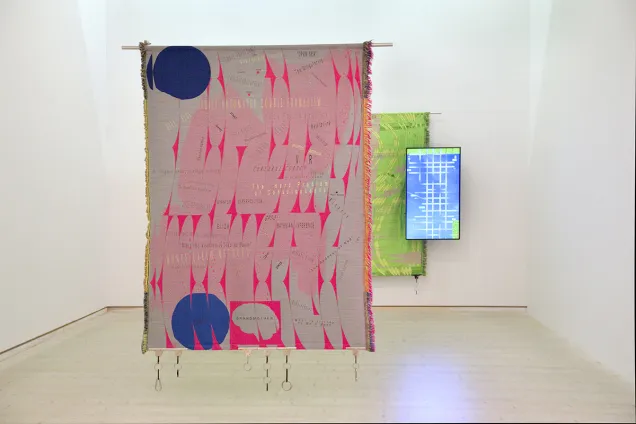 Kodade Trådar 8 Foto Emma Krantz Skissernas Museum .jpg En färgstark väv i rosa, grått och blått hänger från taket i bakgrunden ytterligare en väv, grön, tillsammans med en digital skärm som visar en video.
