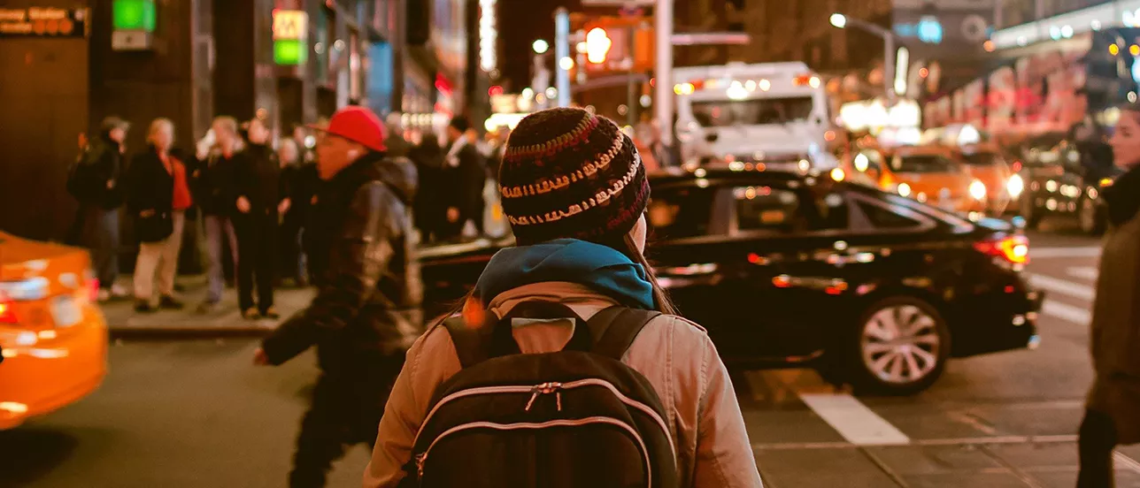 Person med mössa och ryggsäck fotograferad bakifrån i en myllrande stadsmiljö.