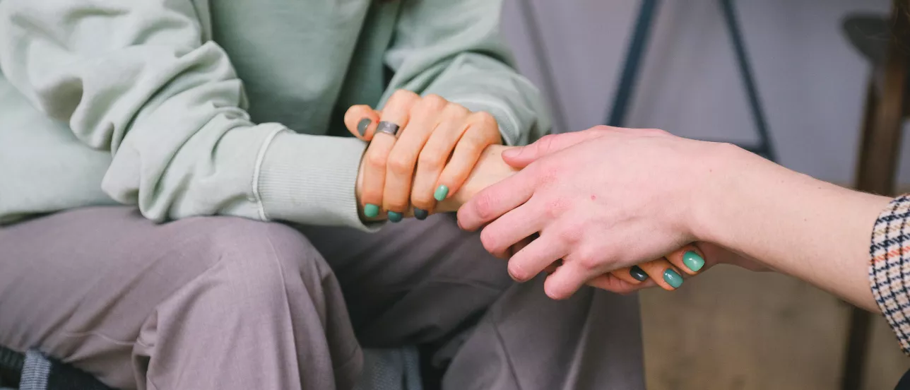 Två personer sitter mitt emot varandra och håller hand. Foto.